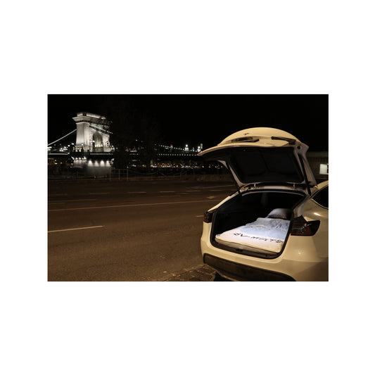 White Tesla Model Y parked at night on a riverside road in Budapest, tailgate open with the EV-MATS mattress made up as a bed inside, the illuminated Chain Bridge in the background.