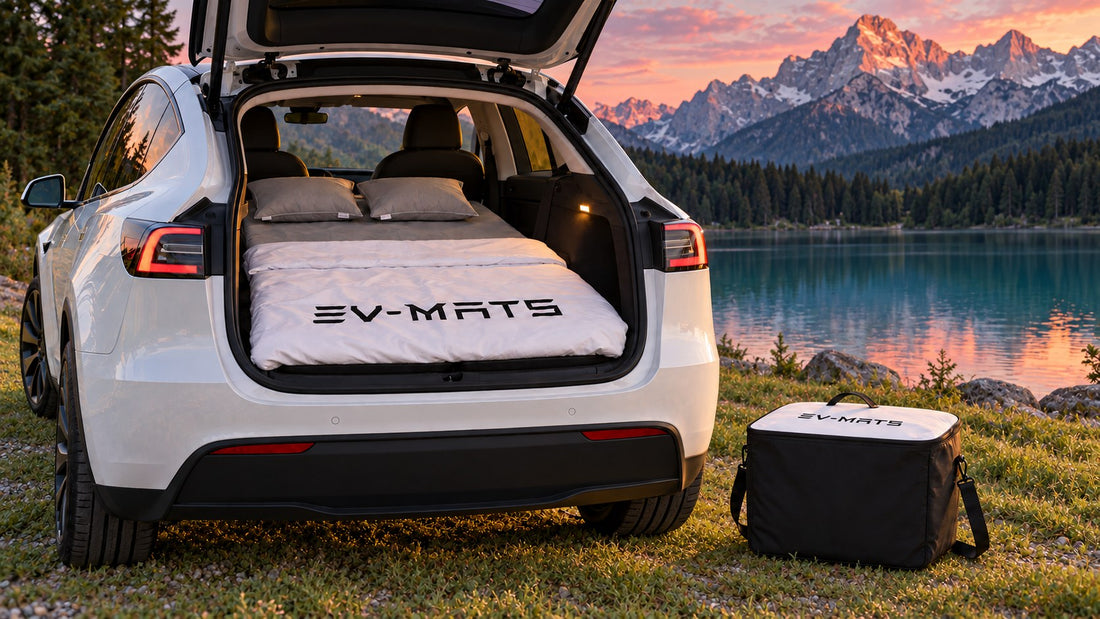 White Tesla Model Y by an Alpine lake at dusk, tailgate open with an EV-MATS bed inside: grey sheet, two grey pillows, white duvet with black EV-MATS logo. The EV-MATS carrying case with white logo lid stands on the grass.